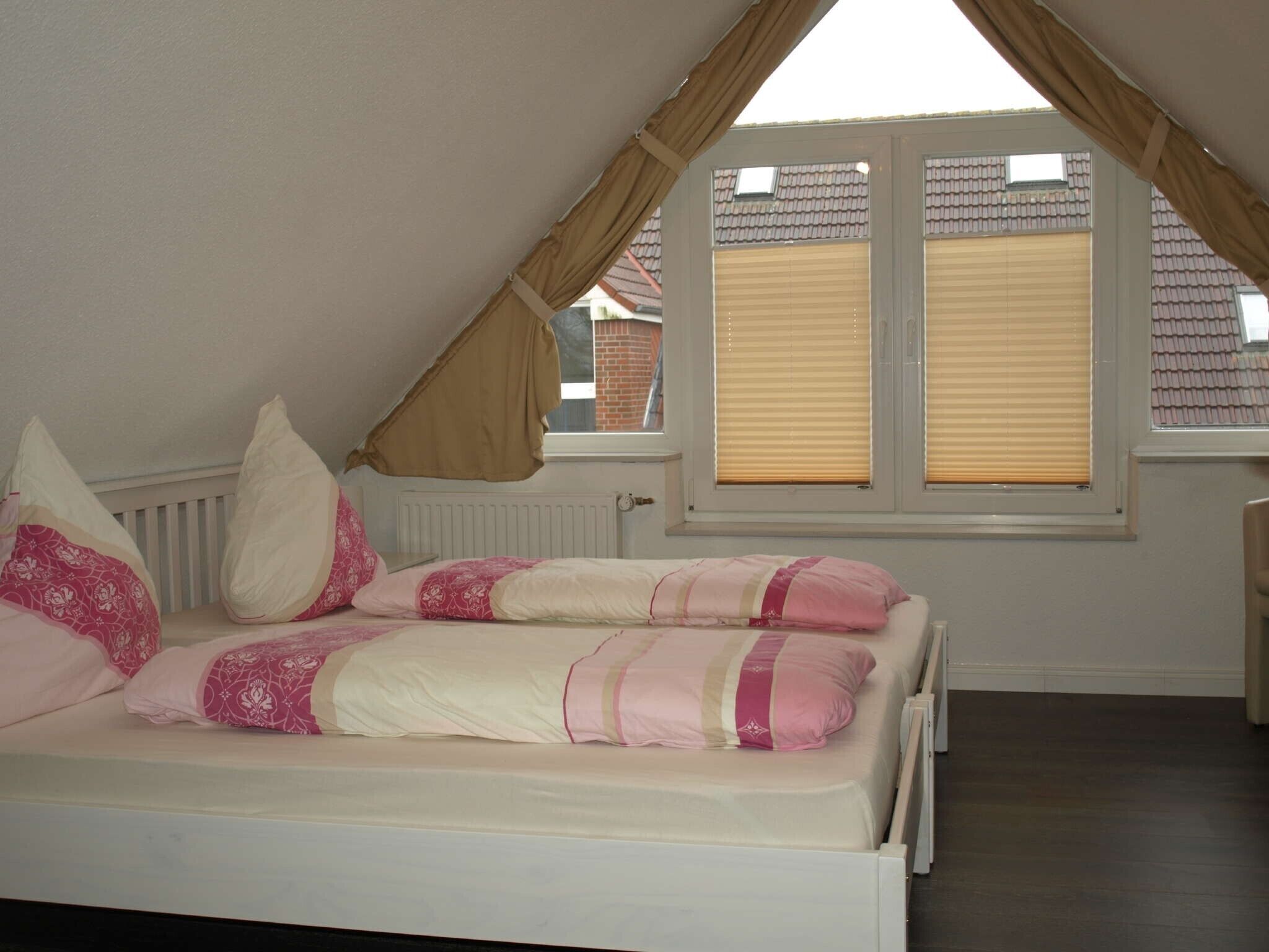 Et soveværelse med skråvæg, seng med pink og hvidt sengetøj, to vinduer med persienner og en radiator.