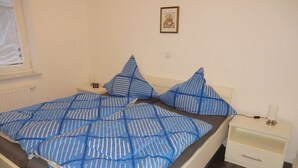 Ein Bett mit blauer Streifenauflage, zwei Kissen, ein Nachttisch mit Lampe und eine kleine Schublade.
