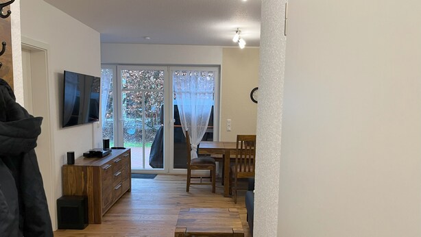 Ein Wohnzimmer mit Fernseher, Holzmöbeln, einer Schiebeglastür, die zu einem Garten führt, und einem Kleiderständer.