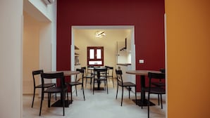 Een eetgedeelte met meerdere tafels en stoelen, een rode accentmuur, en een keuken zichtbaar door een open doorgang.