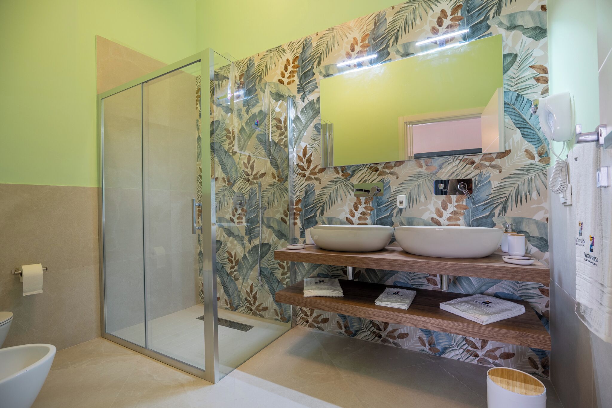 A bathroom with a glass shower enclosure, a large mirror, two sinks with a bowl sink, and a wooden shelf with towels.