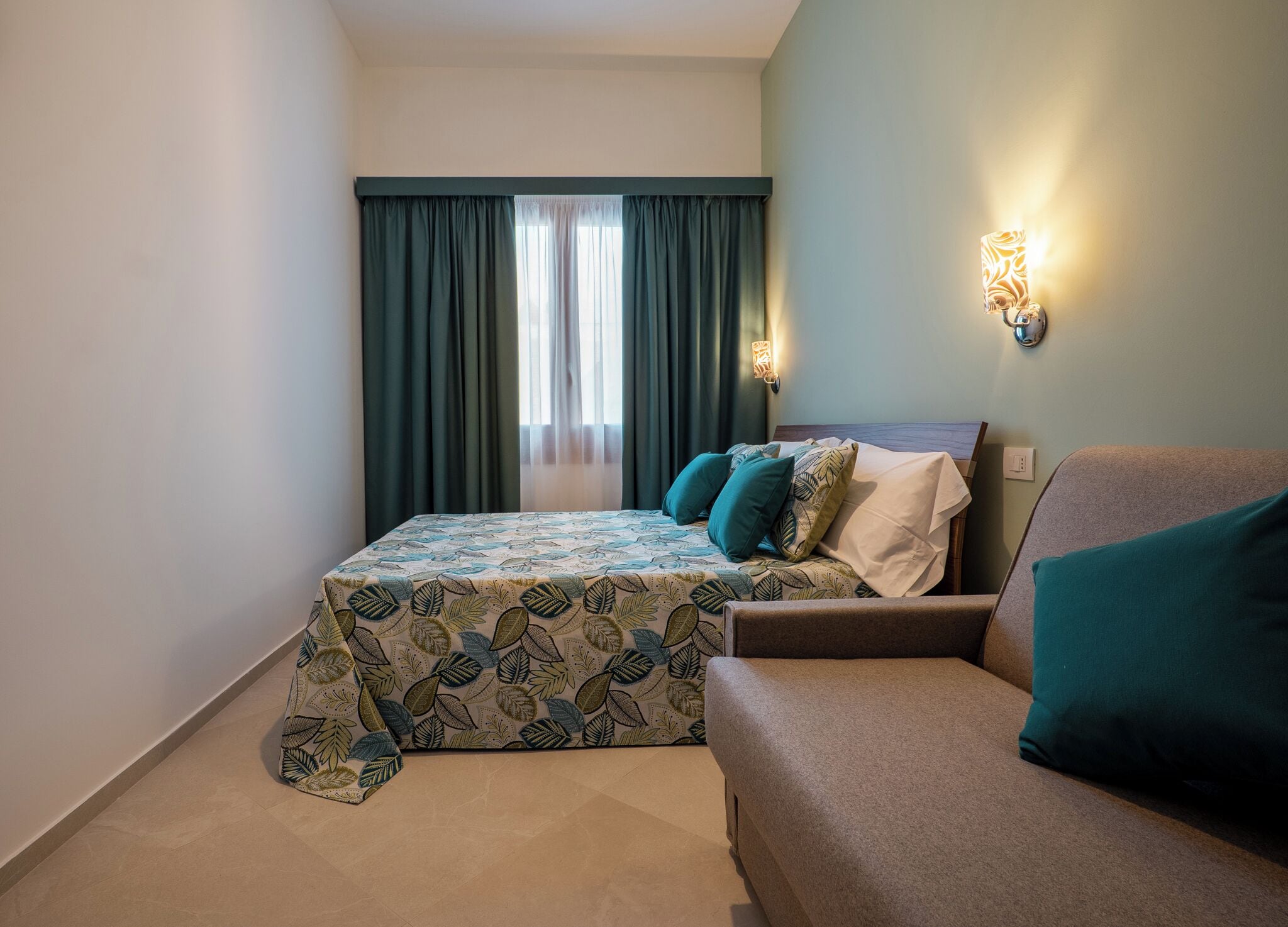 A bedroom with a bed, a sofa, a window with curtains, and wall-mounted lights.