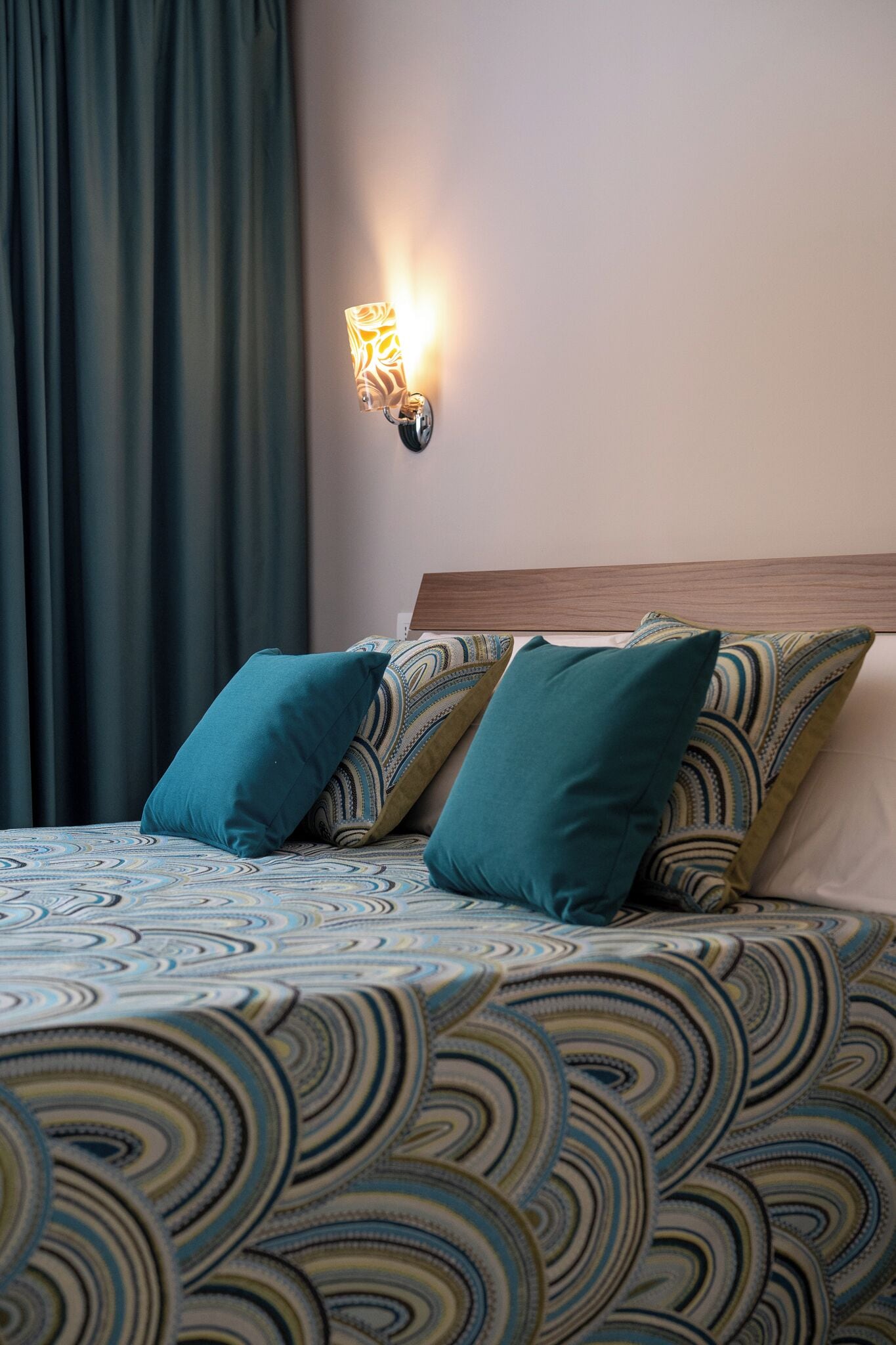 Una cama con ropa de cama estampada, una lámpara fijada a la pared y una cortina oscura.
