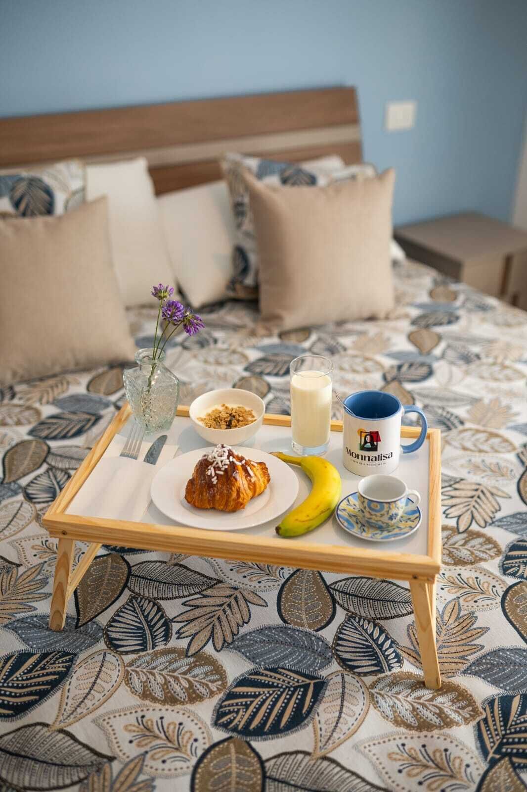Una bandeja de desayuno sobre una cama con una colcha estampada con motivos florales, un tazón de cereal, un croissant, un plátano, un vaso de leche y una taza de café.