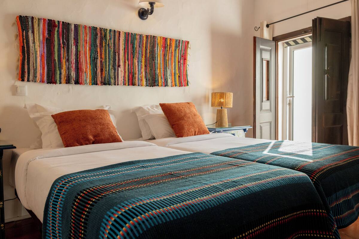 A hotel room with two beds, a colorful tapestry on the wall, and a door to the right.