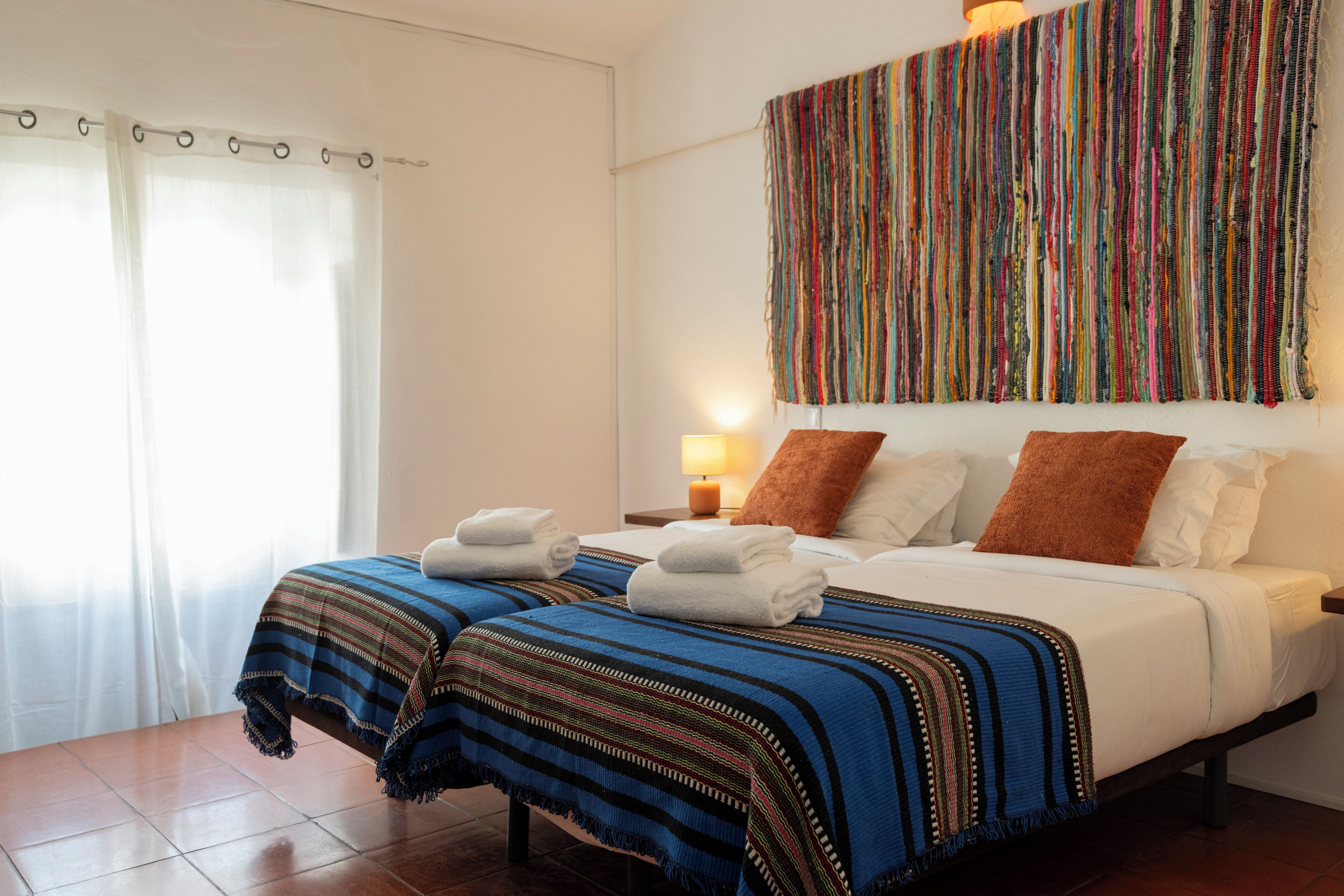 A hotel room with two beds, a colorful tapestry, a nightstand with a lamp, and a window with curtains.