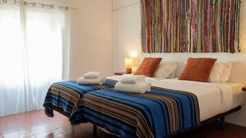A hotel room with two beds, a colorful tapestry, a nightstand with a lamp, and a window with curtains.