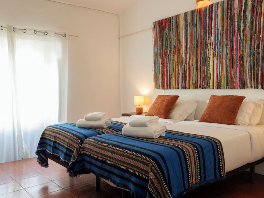 Ein Hotelzimmer mit zwei Betten, einem farbenfrohen Wandteppich, einem Nachttisch mit Lampe und einem Fenster mit Vorhängen.