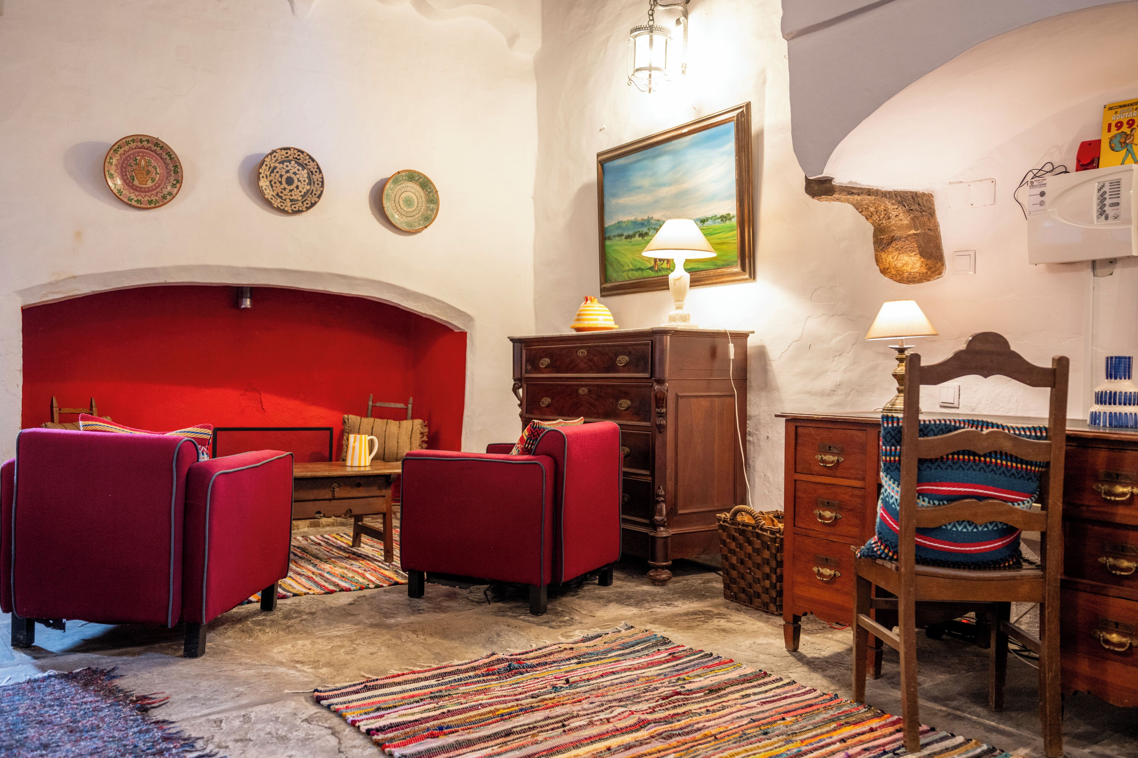 A room with a red sofa, a wooden chair, a small table, a lamp, and a painting on the wall.