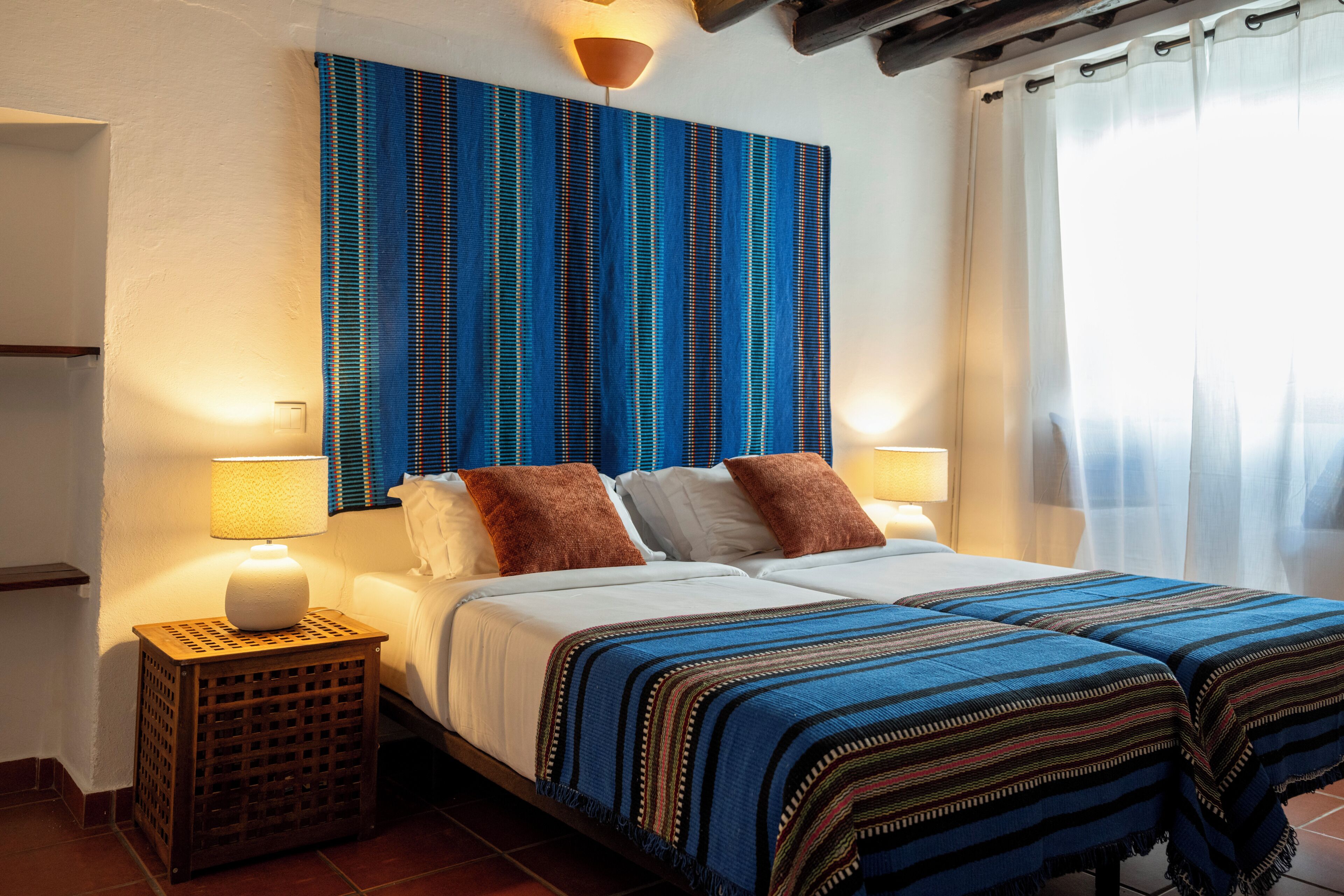 A double bed with two pillows, a nightstand with a lamp, and a blue-striped wall hanging.
