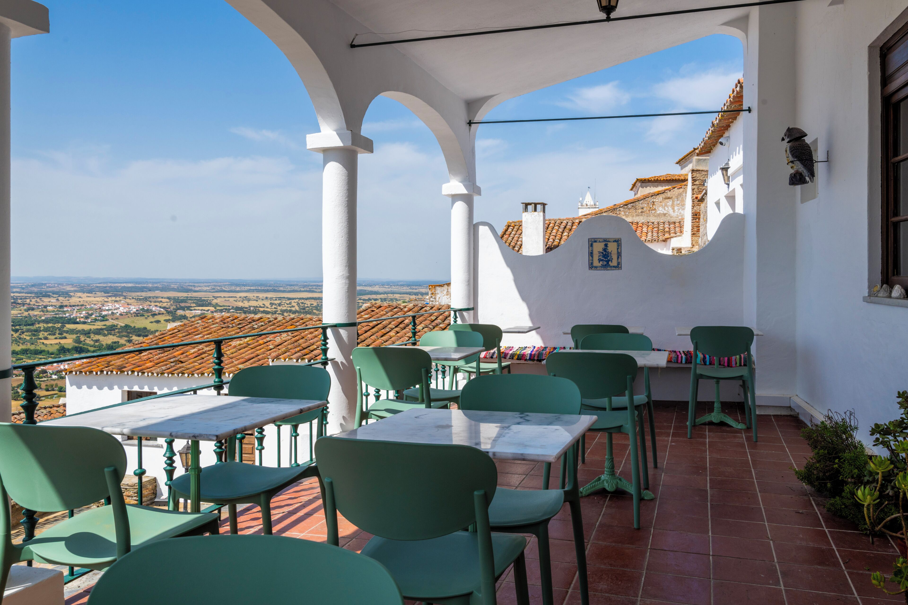 A terrace with green chairs and tables, white walls, and a view of a village.