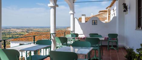 A terrace with green chairs and tables, white walls, and a view of a village.