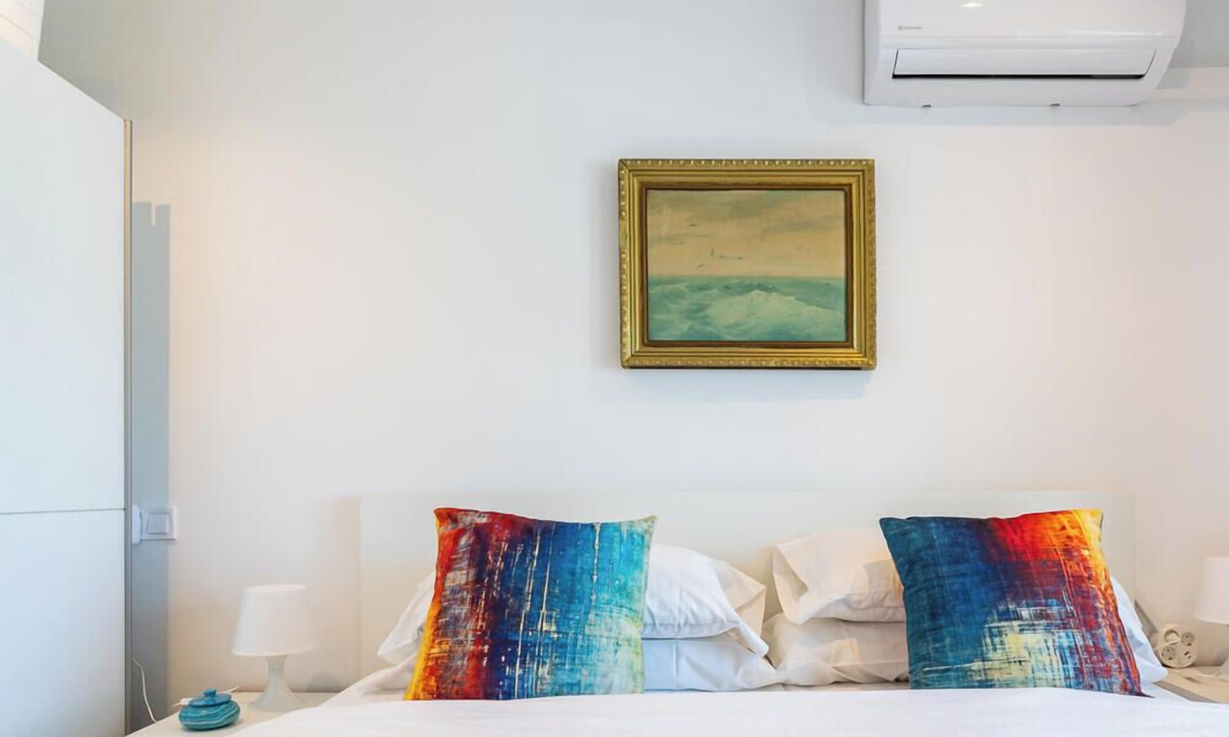 Hotellihuone, jossa on sänky, tyynyjä, taulu ja ilmastointilaite.