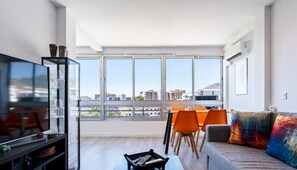 Ein modernes Wohnzimmer mit großem Fernseher, orangefarbenen Sesseln, einer Couch mit bunten Kissen und einem Essbereich mit Blick auf Gebäude.
