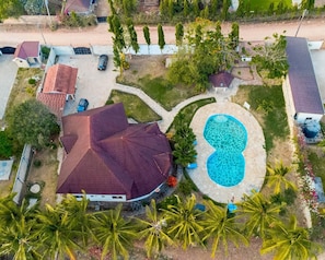 An aerial view of a residential area with a large swimming pool, a house with a red roof, and surrounding greenery.