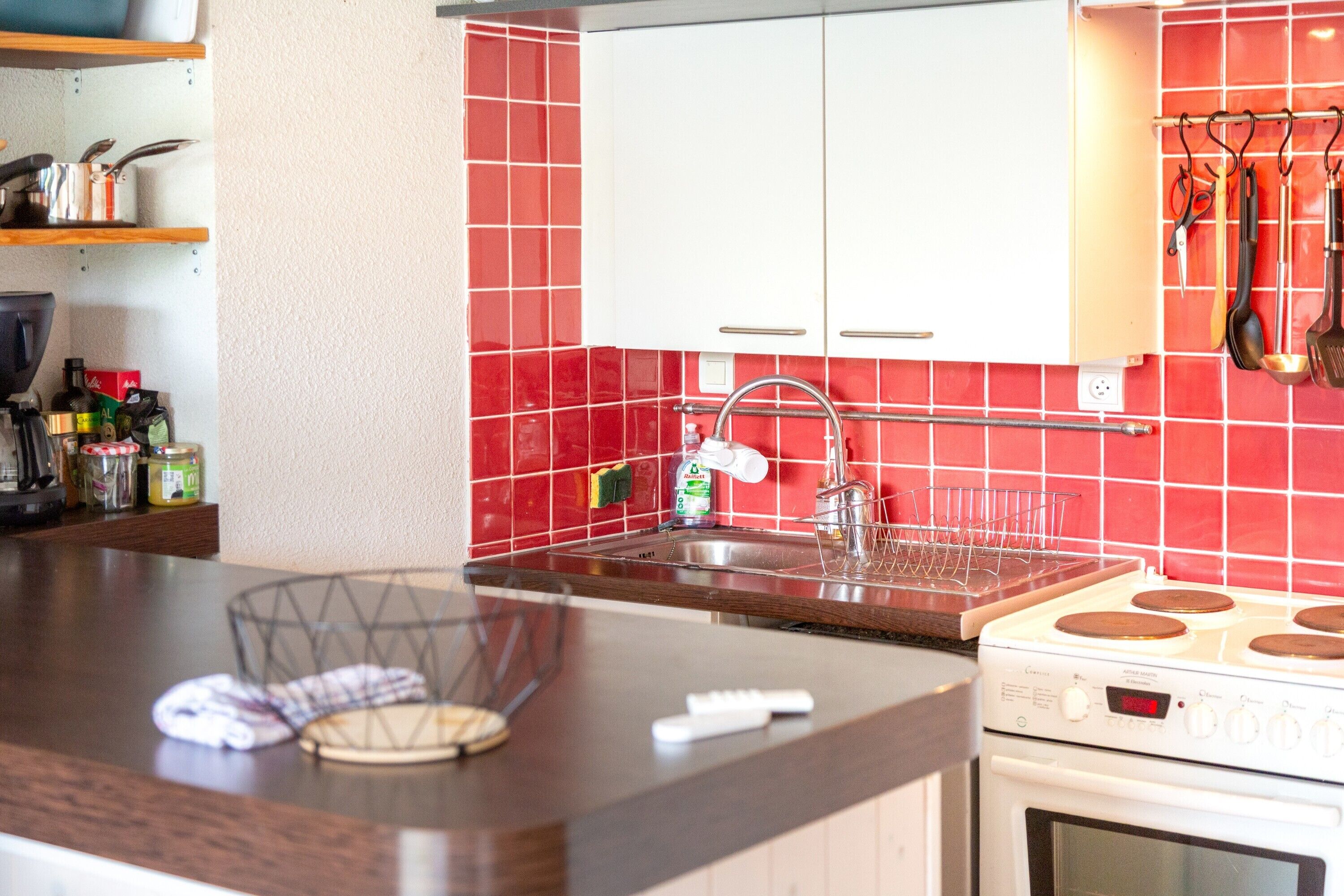 Een keuken met een rode betegelde spatwand, witte kasten, een roestvrijstalen spoelbak en een fornuis.