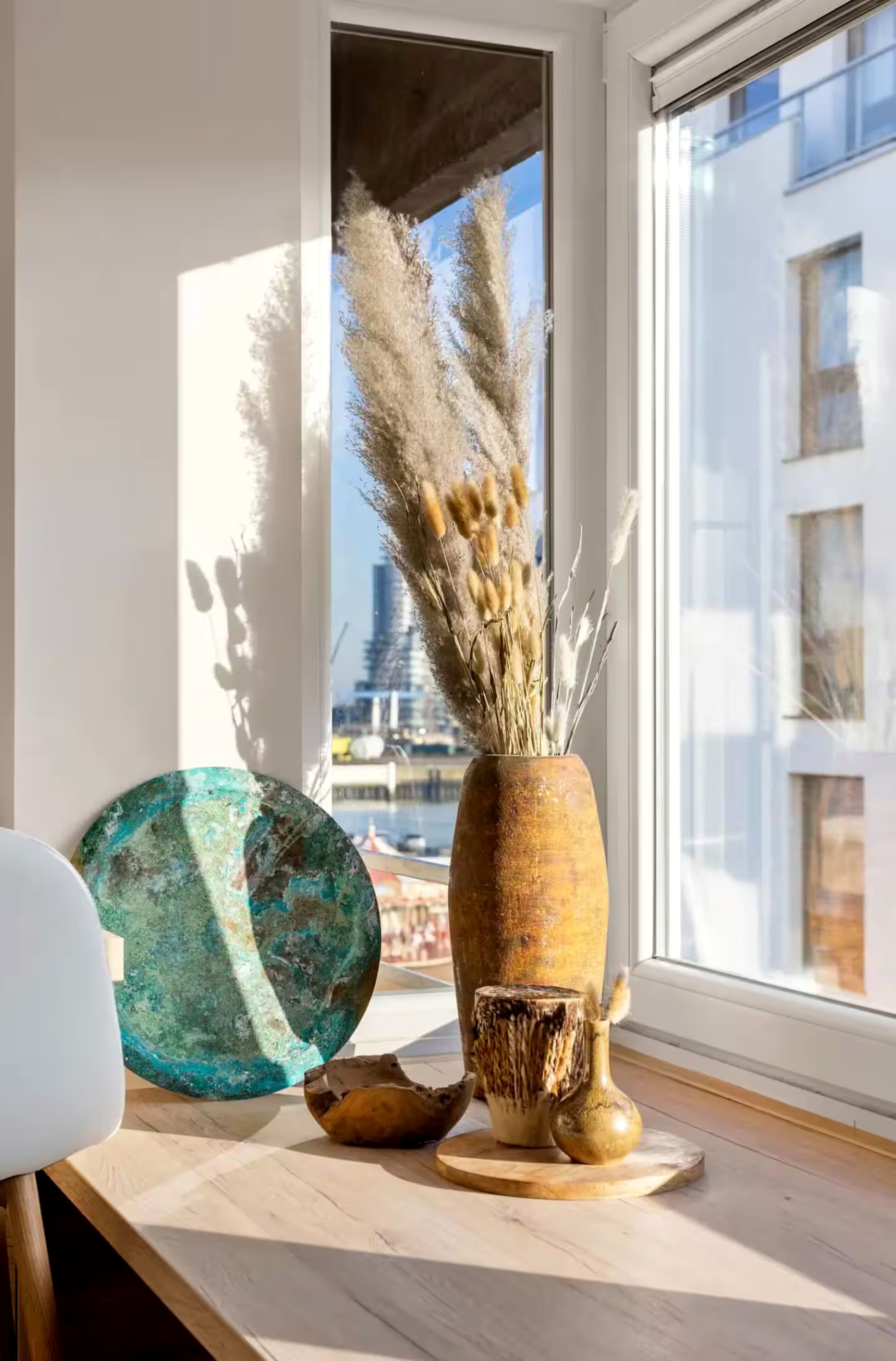 A window with a view of a cityscape, a rustic vase with dried pampas grass, and a teal-colored ceramic piece on a wooden surface.