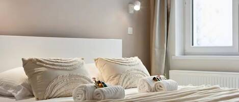 A neatly made bed with pillows and rolled towels, a window with curtains, and a wall-mounted light fixture.