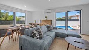 A modern living room with a grey sectional sofa, a dining table with chairs, and a view of a swimming pool through large windows. - Villa Jurić (Vodice)