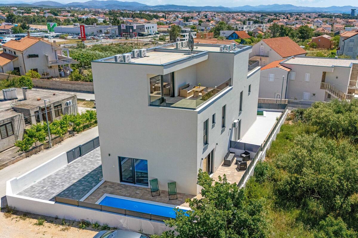 Um edifício moderno de dois andares com piscina no terraço, rodeado por casas residenciais e vegetação.