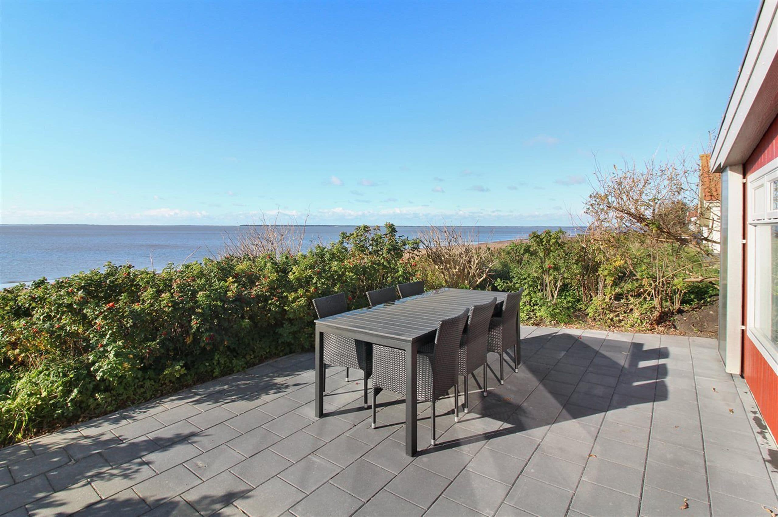 An outdoor patio with a table and chairs, a view of the sea, and a building with a red exterior.
