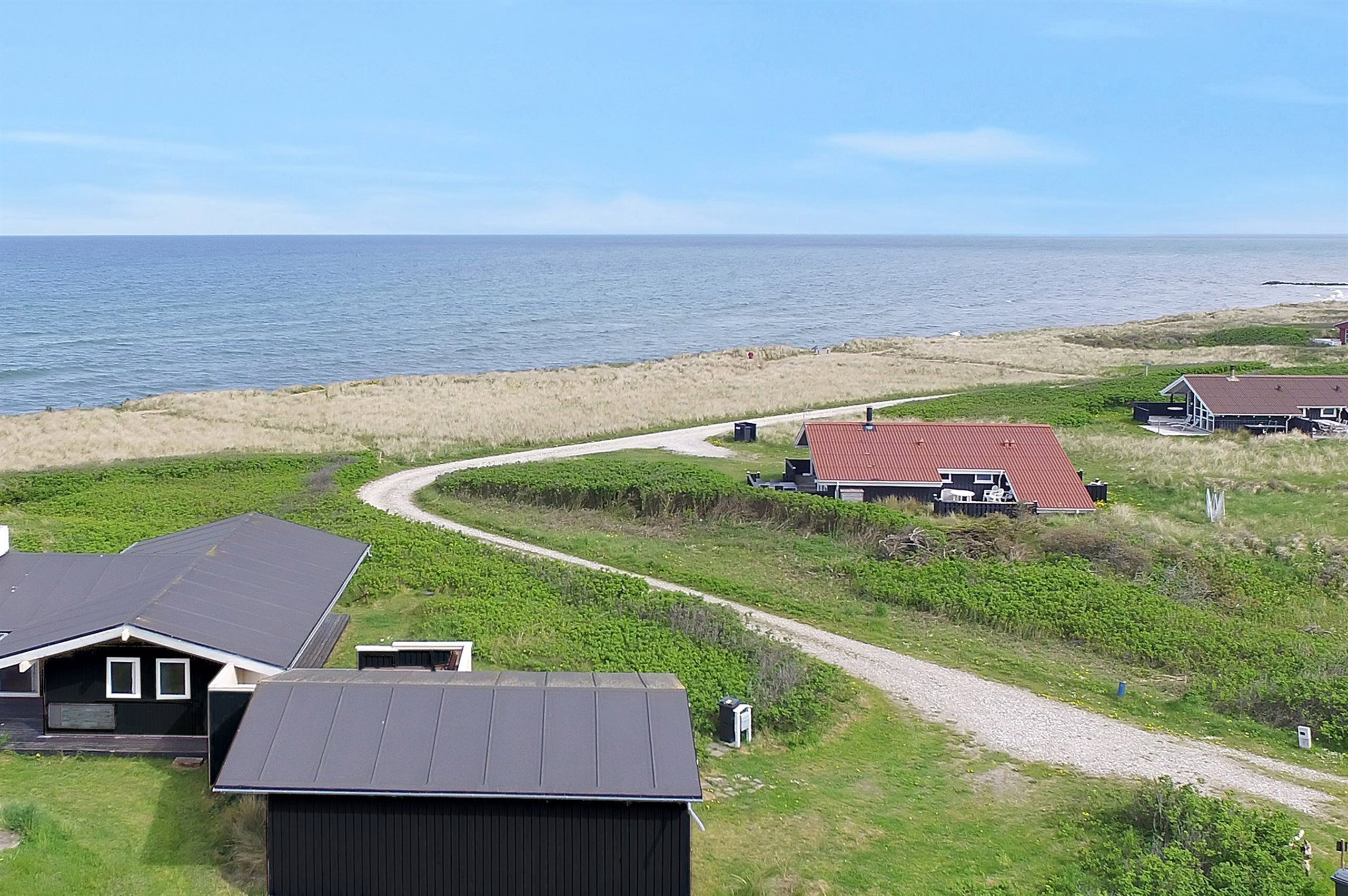 A coastal property with a gravel path leading to a house with a red roof, surrounded by grassy dunes and a view of the sea.