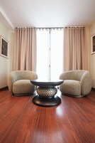 Una habitación con una mesa redonda, dos sillones, piso de madera y cortinas color beige.