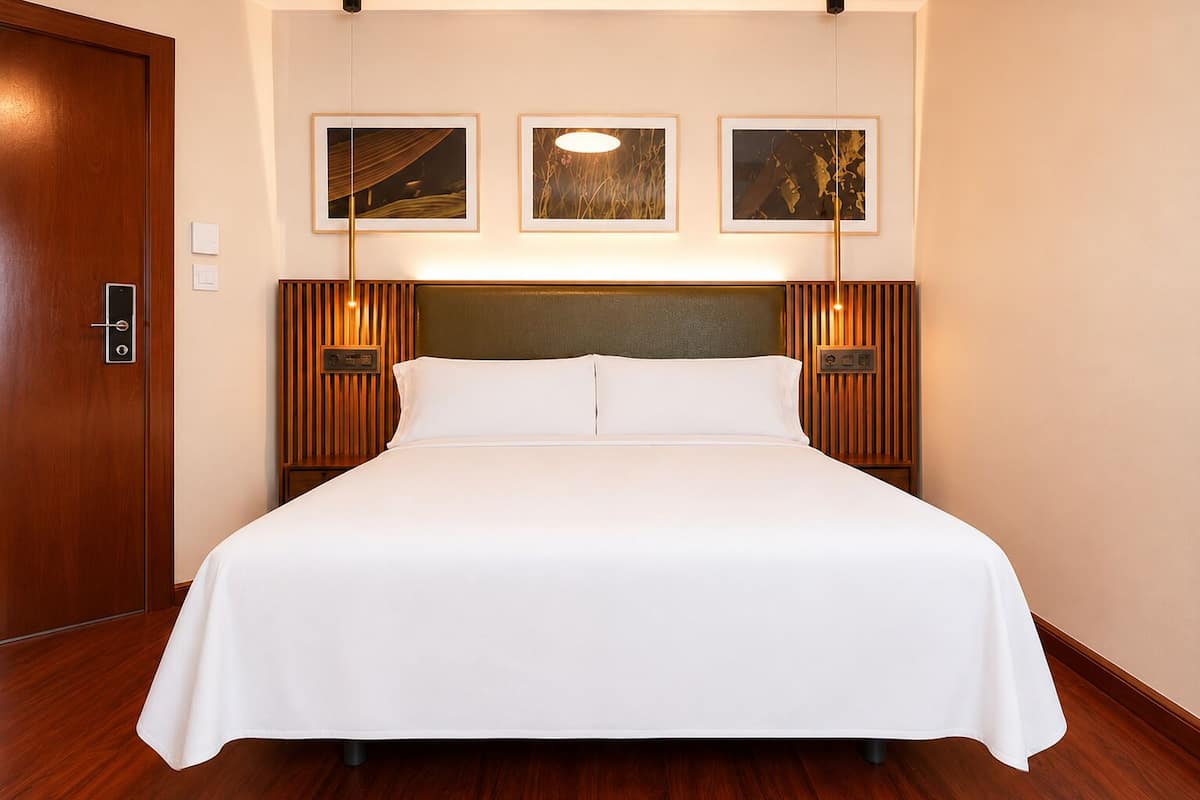 A neatly made bed in a hotel room with a wooden headboard, framed artwork on the wall, and a door with a handle.