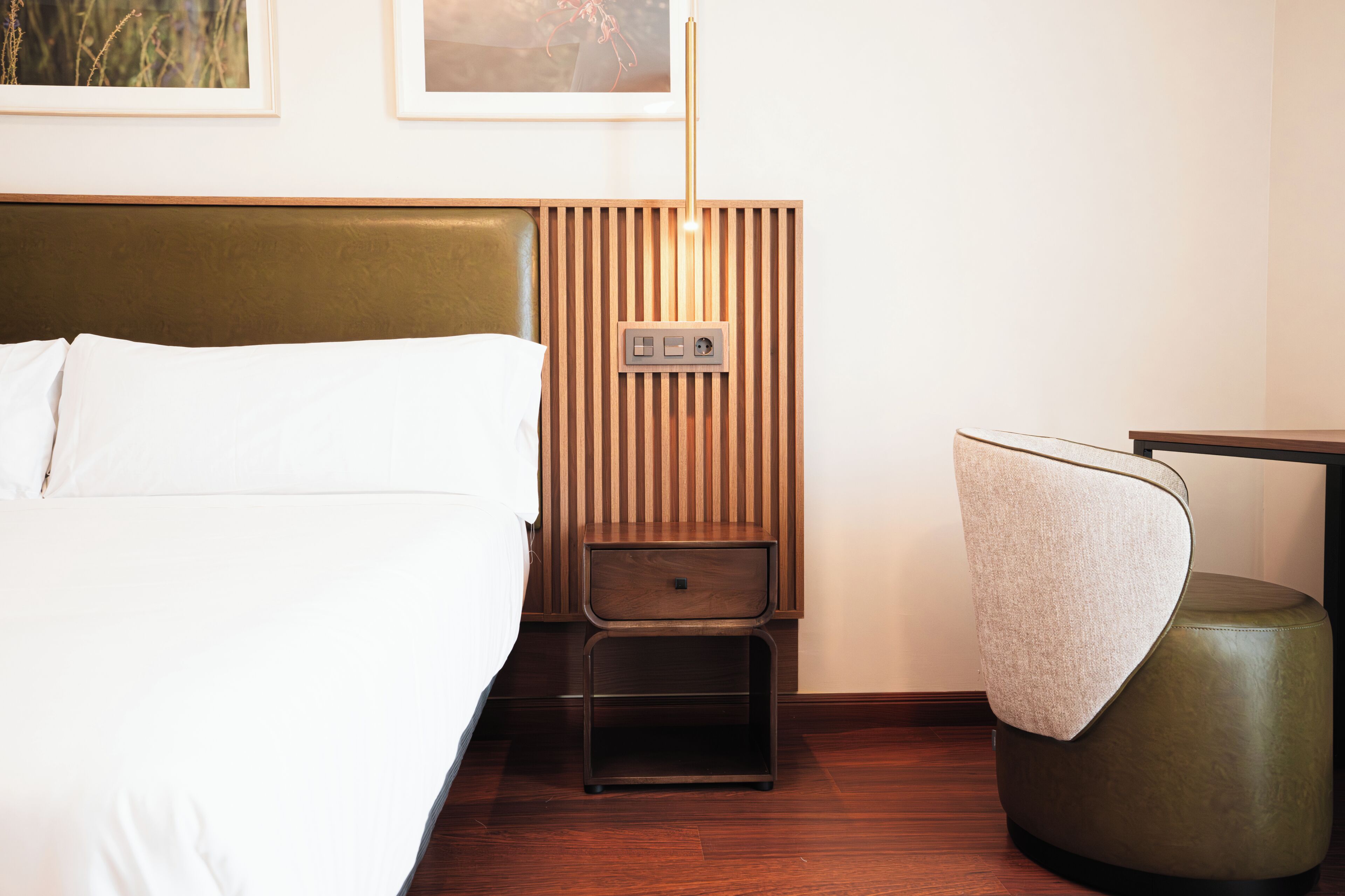 Een hotelkamer met een bed, nachtkastje met lamp, een stoel en een bedieningspaneel aan de muur.
