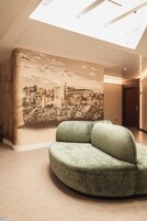 Un interior moderno con un sofá verde curvo, un mural de un paisaje urbano histórico y un tragaluz.
