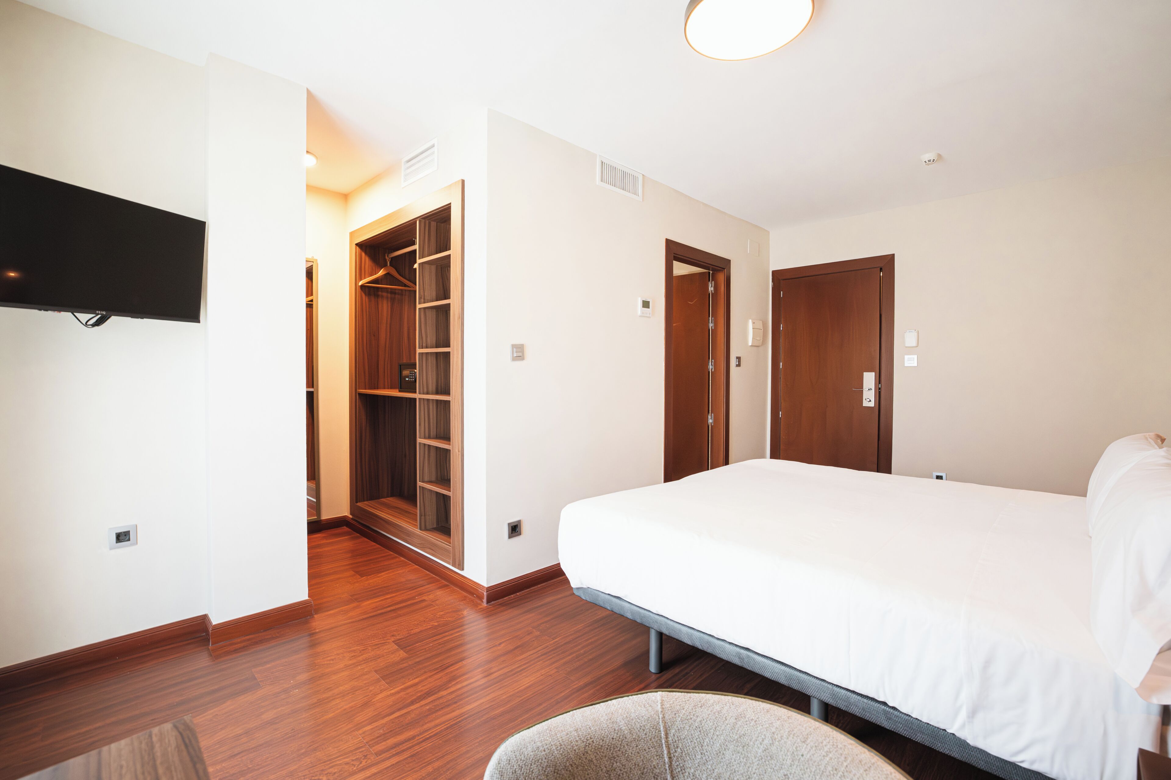 Una habitación de hotel con cama, televisor montado en la pared, piso de madera y vestidor.