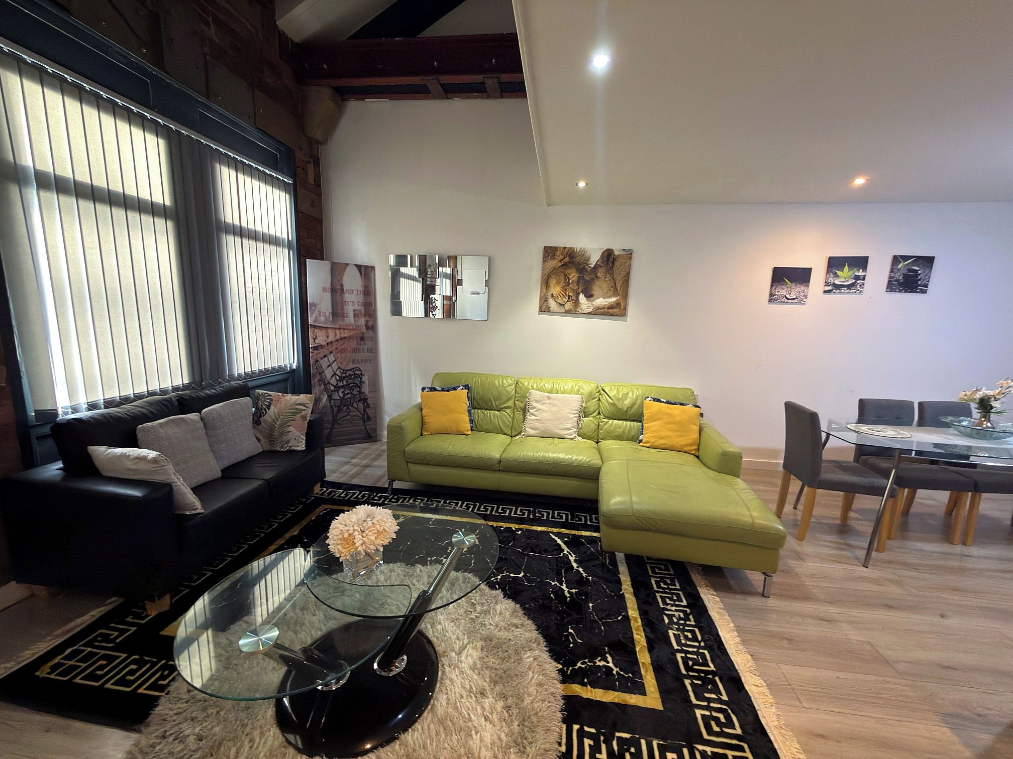 A modern living room with a green sofa, black and green patterned rug, and a glass coffee table with a flower vase.