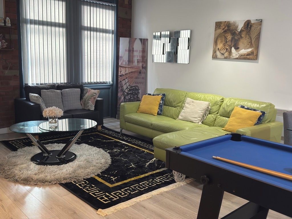 A living room with a green sofa, a pool table, a round coffee table with a vase, and a large window with blinds.