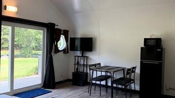 Un espacio habitable compacto con zona de comedor, televisor de pantalla plana, refrigerador negro y un amplio ventanal corredero que da a un área exterior.
