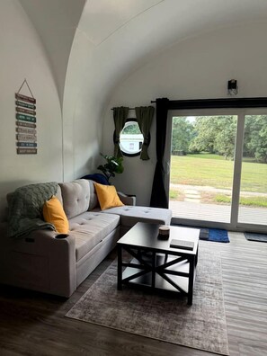 A modern living room with a sofa, a coffee table, and a view through a large sliding glass door to an outdoor area. - Charming Ranch Retreat with Mini Goats & Paddle Boats near Littleton, NC (Littleton)