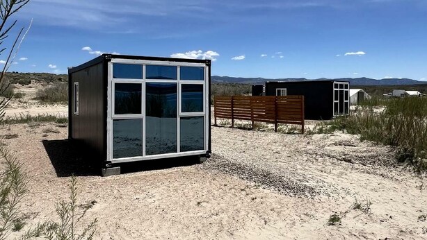 A small, modern building with a large windowed front, a bench, and a dirt ground.