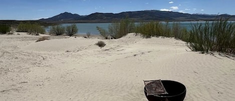 A desert landscape with a large body of water, mountains in the distance, and a black bowl on the sand.