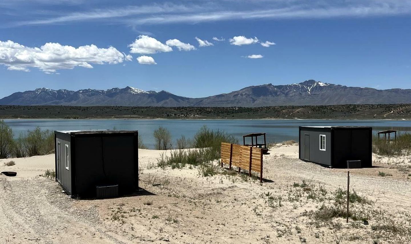 A desert landscape with two black cabins, a wooden bench, and a mountain range in the background.