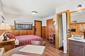 Ein Schlafzimmer mit Bett, Nachttisch, Lampe, Fenster und einem Badezimmer mit Waschbecken und Spiegel.