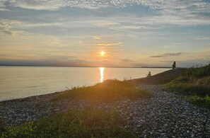 Ein Sonnenuntergang über ruhigem Gewässer mit steiniger Küste, eine Person sitzt am Ufer.