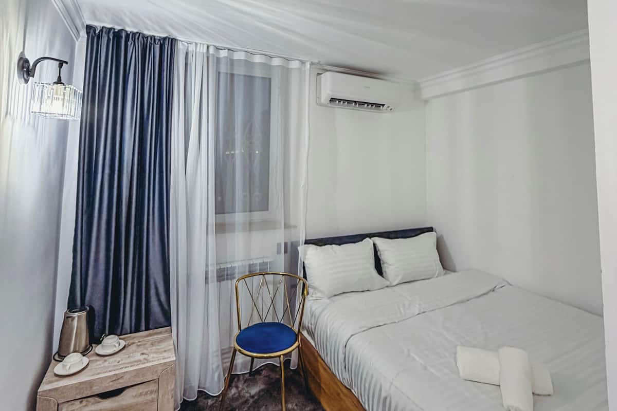 Hotellihuone, jossa on sänky, tuoli, yöpöytä, ilmastointilaite ja ikkuna, jossa on verhot.