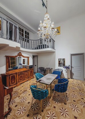 Ruime kamer met een kroonluchter, een eethoek met blauwe gestoffeerde stoelen, een houten kast en een vloer met patroon.