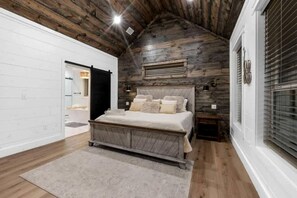 A bedroom with a wooden ceiling, a bed with multiple pillows, a nightstand, and a window with blinds. - Luxury Cabin with Private Hot Tub, Infrared Sauna & Vintage Jukebox Near Broken Bow, OK (Broken Bow)