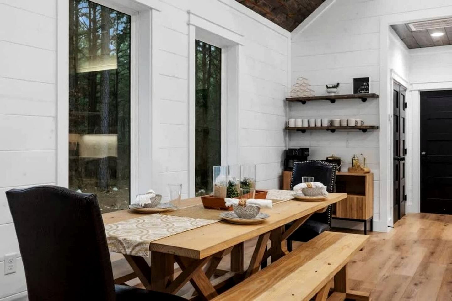 A dining area with a wooden table set for four, surrounded by chairs. There are shelves above the table holding cups and a sign.