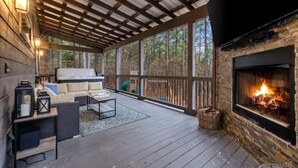 A covered deck with a fireplace, outdoor seating, and a view of the surrounding forest. - Luxury Escape Perfect for Families with Private Hot Tub, Infrared Sauna & Vintage Jukebox in  Broken Bow, Oklahoma (Broken Bow)