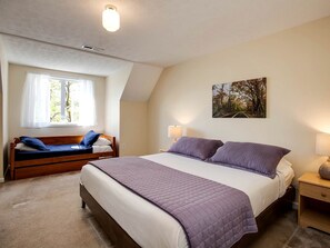 A bedroom with a large bed, a smaller bed, a window with curtains, a painting on the wall, and a bedside table with a lamp.