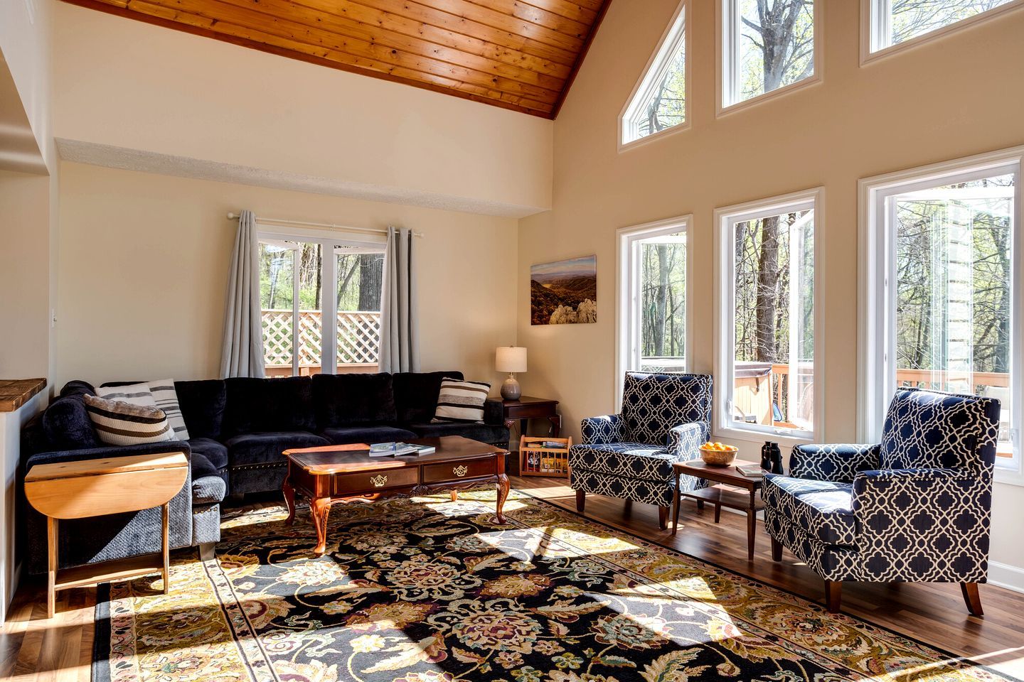 Un salon spacieux avec un plafond haut, de grandes fenêtres, un canapé de couleur foncée, des fauteuils à motifs et un tapis décoratif.