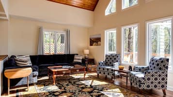 A spacious living room with a high ceiling, large windows, a dark-colored sofa, patterned armchairs, and a decorative rug.