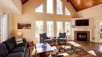 A spacious living room with a fireplace, large windows, and a high ceiling.