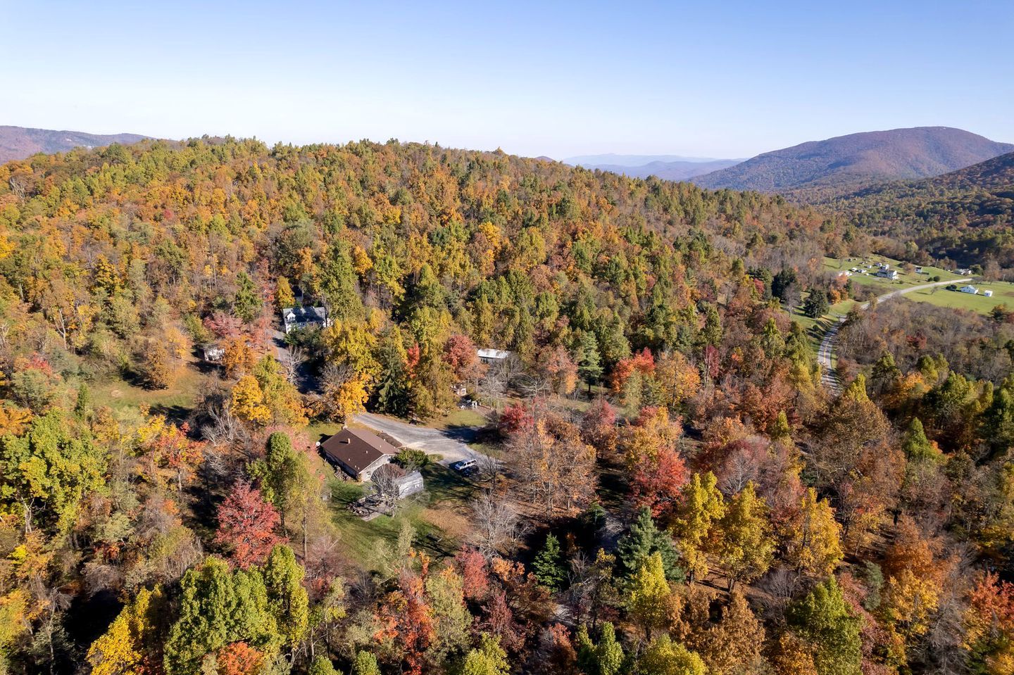 A residential area with houses surrounded by dense forest, a winding road, and distant mountains.
